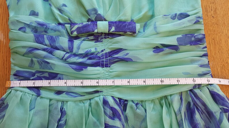 May include: Close-up of a light green dress with blue floral patterns. The dress features a bow at the waist and a gathered bodice. A measuring tape shows a width of approximately 13 inches.