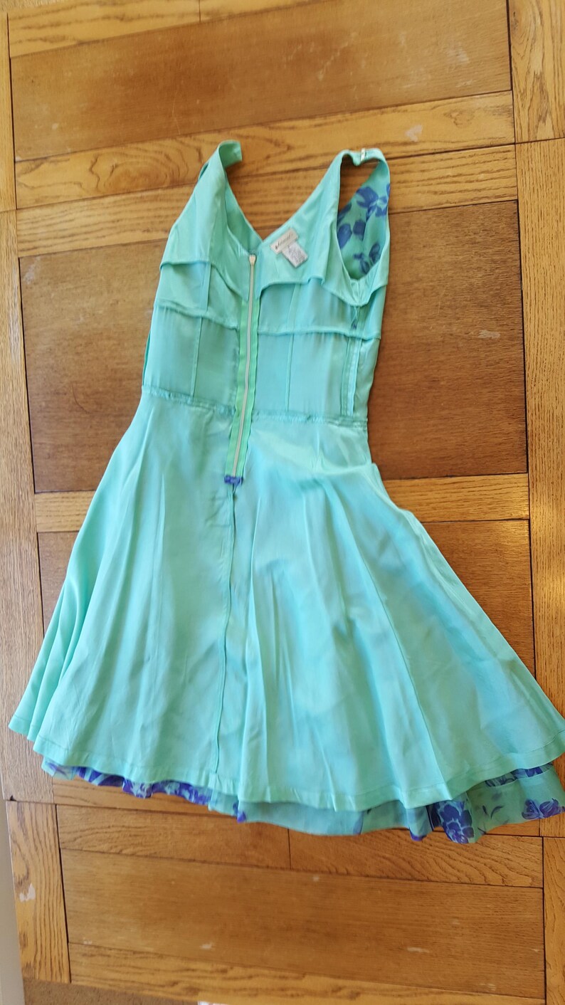 May include: A light turquoise dress with a V-neckline and a full skirt. The dress features a front zipper and a contrasting floral pattern on the lining. The dress is made of a lightweight fabric, perfect for warm weather.
