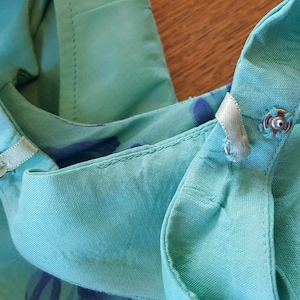 May include: Close-up of a turquoise top with a halter neckline. The fabric has a subtle floral pattern in shades of blue and purple. The top features a snap closure and delicate ribbon details.