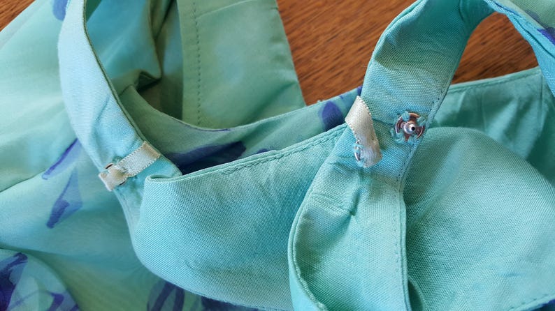 May include: Close-up of a turquoise top with a halter neckline. The fabric has a subtle floral pattern in shades of blue and purple. The top features a snap closure and delicate ribbon details.