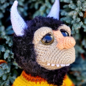 May include: A handmade crochet monster doll with large, reflective eyes, a tan muzzle, and white horns. The doll has a black fur mane and is wearing a yellow and orange striped sweater. The doll is set against a backdrop of green foliage.