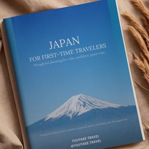 以下が含まれることがあります： 「JAPAN」と書かれた旅行ガイド。表紙には青い空と山の風景が描かれています。「FOR FIRST-TIME TRAVELERS」と「Thoughtful planning for calm, confident Japan trips」と書かれています。初めての日本旅行を安心して楽しめるように設計されています。