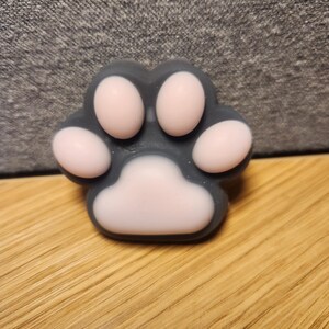 May include: A gray and pink paw-shaped soap. The soap has a dark gray outline with pink pads. The soap is sitting on a wooden surface.