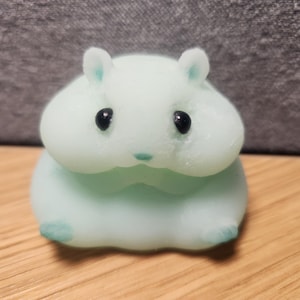 May include: A light blue hamster figurine with black eyes and small ears. The figurine is sitting on a wooden surface. The hamster has a cute, rounded shape and is a light blue color with darker blue accents on its ears and feet.