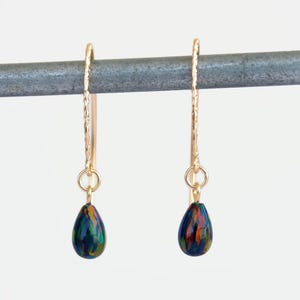 May include: A pair of gold-colored drop earrings. Each earring features a long, textured hook and a teardrop-shaped bead with a mix of blue, green, and orange colors. The earrings hang against a white background.