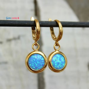 May include: A pair of gold hoop earrings with a dangling blue opal stone. The earrings are on a black stand.