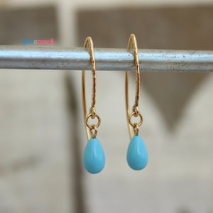 May include: A pair of gold-colored hoop earrings with a hammered texture. Each earring features a small, teardrop-shaped, light blue bead suspended from a delicate chain. The earrings are displayed against a blurred background.