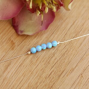 May include: A delicate necklace featuring six light blue opal beads strung on a thin silver chain. The beads are evenly spaced and have a subtle iridescent quality. The necklace is displayed on a wooden surface, with a blurred pink and yellow flower in the background.