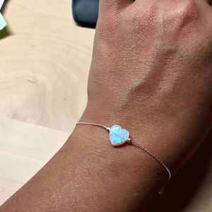 May include: A delicate silver chain bracelet featuring a heart-shaped opal gemstone. The opal displays iridescent hues of blue, green, and pink.