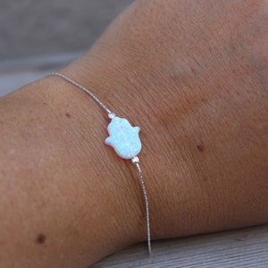 May include: A delicate silver bracelet featuring a Hamsa hand charm. The charm is a light blue opal colour. The bracelet is worn on a wrist.