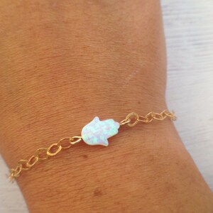 May include: A delicate gold-coloured chain bracelet with a white opal Hamsa hand charm. The bracelet has a textured chain and a shimmering, iridescent charm. The Hamsa hand is a symbol of protection and good fortune.