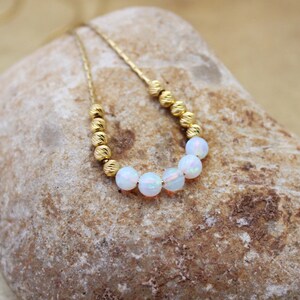 May include: A delicate gold chain necklace with a row of white opal beads. The necklace is displayed on a light brown rock.