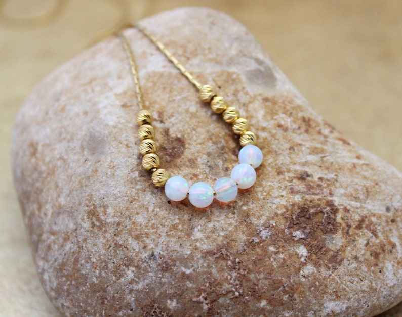 May include: A delicate gold chain necklace with a row of white opal beads. The necklace is displayed on a light brown rock.