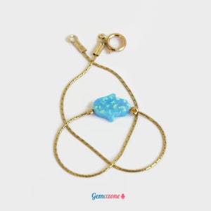 Puede incluir: Una pulsera de cadena dorada con un dije de mano de Hamsa de ópalo azul claro. La pulsera tiene un diseño de cadena delicada y un cierre de mosquetón. El dije es de color azul claro con una superficie texturizada.