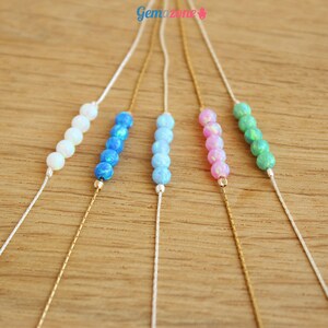 May include: Five delicate necklaces with a silver chain and a row of five opal beads. The necklaces are displayed on a wooden surface. The beads are white, light blue, blue, pink, and green.
