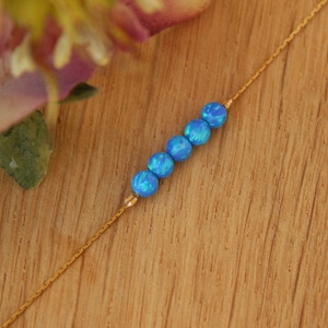 May include: A gold necklace with six blue opal beads. The beads are strung on a fine gold chain. The necklace is displayed on a wooden surface. The opal beads are approximately 0.6 cm in diameter.