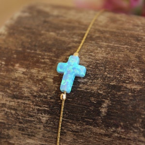Puede incluir: Un delicado brazalete de cadena dorada con un dije de cruz de ópalo azul. La cruz es de color azul claro con un acabado brillante e iridiscente.
