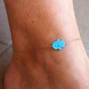 Könnte beinhalten: Ein goldfarbenes Kettenarmband mit einem blauen Opal-Hamsa-Hand-Anhänger. Die Hamsa-Hand ist ein Schutzsymbol. Das Armband wird am Knöchel getragen.