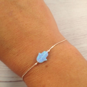 May include: A delicate silver bracelet featuring a light blue opal Hamsa hand charm. The bracelet is worn on a wrist, showcasing the charm's intricate details and subtle color variations. The Hamsa hand is a symbol of protection and good fortune.