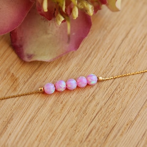 May include: A delicate gold chain necklace with a row of six pink opal beads. The necklace is displayed on a wooden surface.