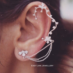 May include: Silver ear cuff with a delicate floral design and a dangling chain. The ear cuff is worn on the upper part of the ear and has a small flower stud earring in the lower lobe.