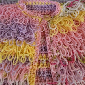 May include: A pastel-coloured, handmade crochet cardigan. The garment features a textured design with looped yarn details and a wooden button closure. The colours include pink, yellow, and lavender. The style is soft and whimsical.