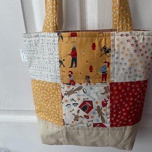 May include: A patchwork tote bag with various dog-themed fabric panels. The bag features a beige base and handles, with panels showcasing dog bones, paw prints, and dog-walking scenes. The word "WOOF" is printed on some panels.