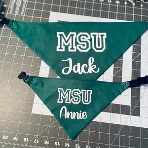 May include: Two green triangular dog bandanas with white text. The top bandana reads "MSU Jack" and the bottom bandana reads "MSU Annie". Each bandana has a black adjustable buckle.