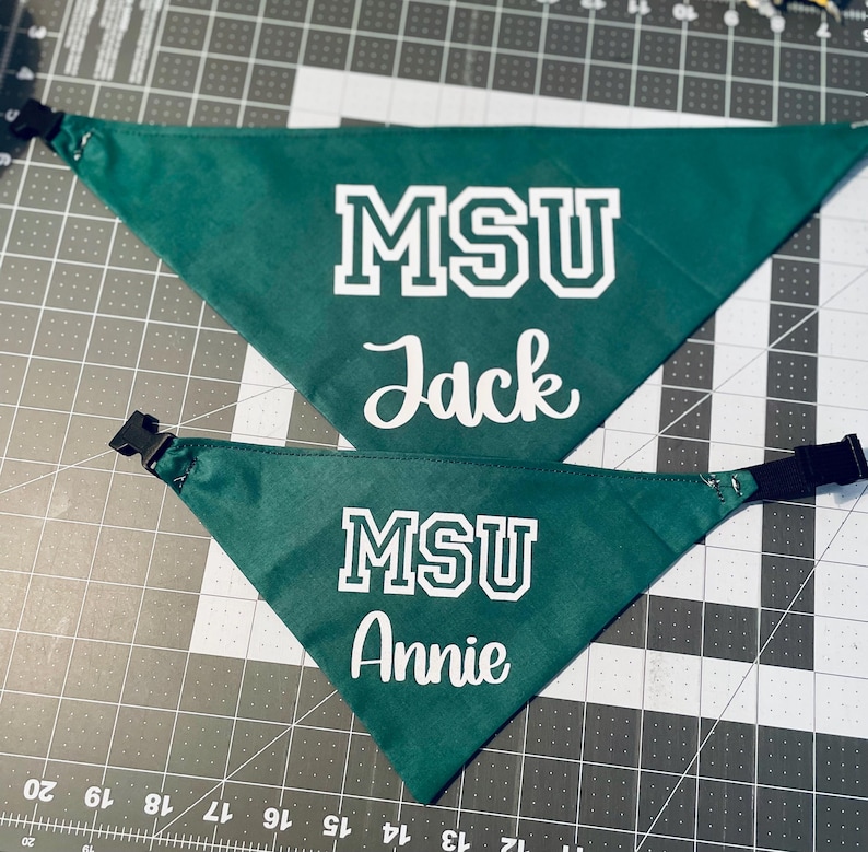 May include: Two green triangular dog bandanas with white text. The top bandana reads "MSU Jack" and the bottom bandana reads "MSU Annie". Each bandana has a black adjustable buckle.