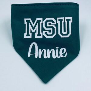 May include: A dark green pet bandana with white text that reads "MSU" in a block letter font and "Annie" in a cursive font. The bandana has a triangular shape and a stitched edge.