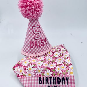 May include: Pink and white gingham party hat with a pink pom pom on top. The hat has the number 5 and the name Daisy embroidered on it. A pink and white floral fabric with the text "Birthday Girl" is underneath the hat.