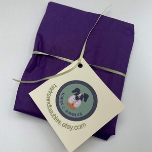 May include: A wrapped gift in deep purple tissue paper, tied with a light green ribbon. A cream-colored tag with a dog illustration and the text "barksandbaubles.etsy.com" is attached.