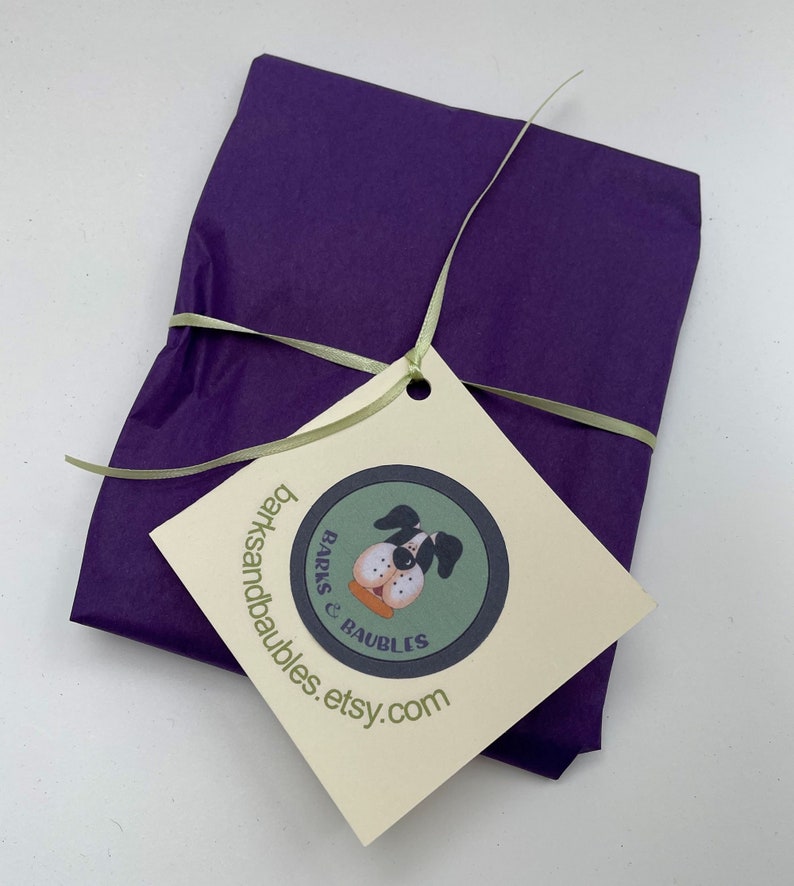 May include: A wrapped gift in deep purple tissue paper, tied with a light green ribbon. A cream-colored tag with a dog illustration and the text "barksandbaubles.etsy.com" is attached.