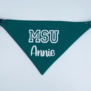 May include: Green dog bandana with the text "MSU" in white block letters and the name "Annie" in white script. The bandana has a black adjustable buckle and strap.