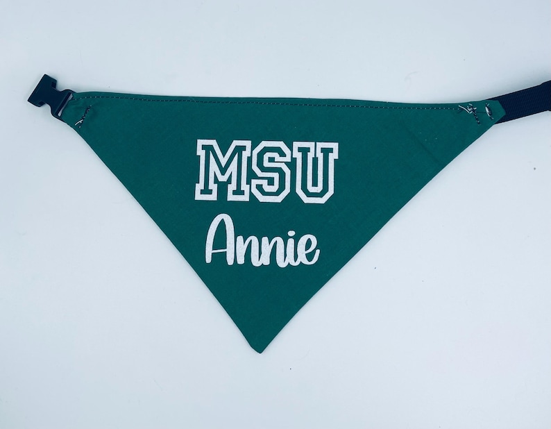 May include: Green dog bandana with the text "MSU" in white block letters and the name "Annie" in white script. The bandana has a black adjustable buckle and strap.