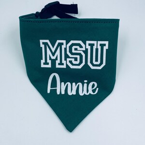 May include: A green pet bandana with the text "MSU Annie" in white block letters and script. The bandana has a black adjustable strap. The fabric is a solid green color.