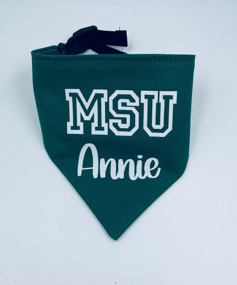 May include: A green pet bandana with the text "MSU Annie" in white block letters and script. The bandana has a black adjustable strap. The fabric is a solid green color.