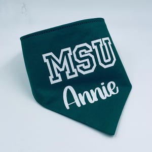 May include: A dark green pet bandana with white text. The bandana features the letters "MSU" in a block font and the name "Annie" in a cursive font. The edges are stitched with a dark thread.