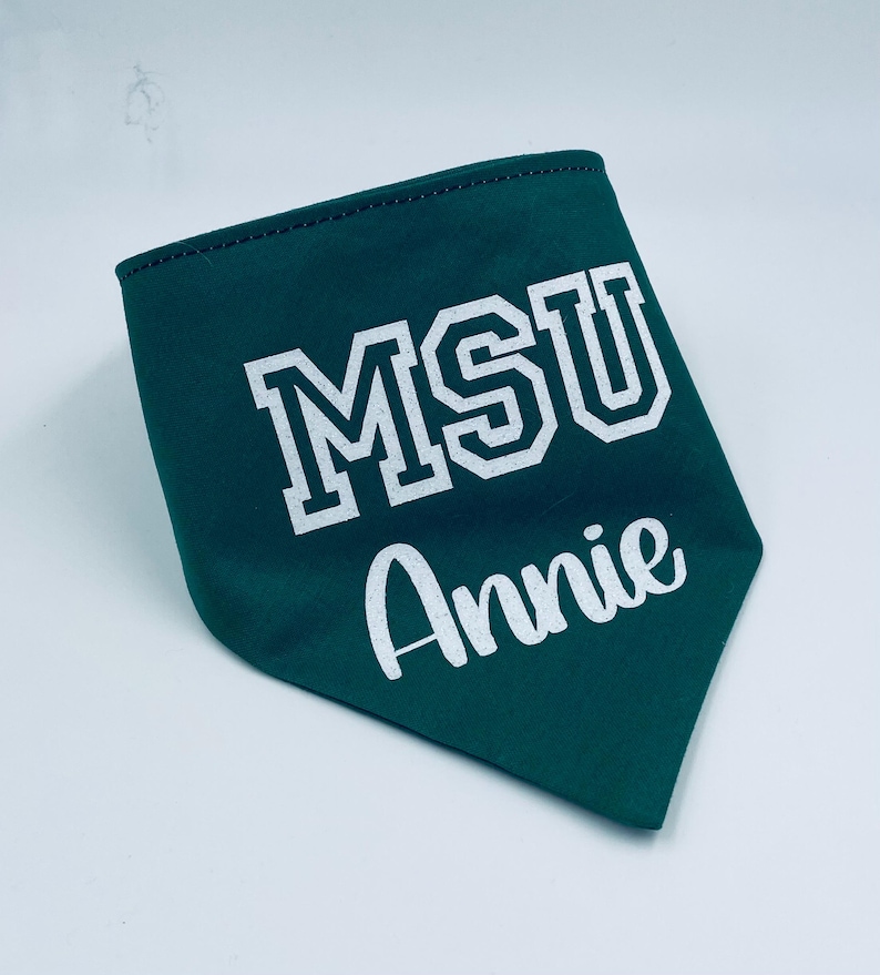 May include: A dark green pet bandana with white text. The bandana features the letters "MSU" in a block font and the name "Annie" in a cursive font. The edges are stitched with a dark thread.