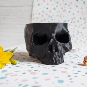 May include: A black skull-shaped planter with a small gold skull figurine on a white background with colourful flecks.