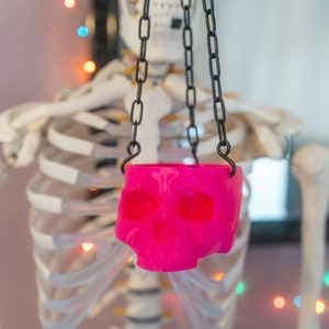 May include: A bright pink skull-shaped planter suspended by black chains. The planter has eye and nose sockets, and a mouth. The background features a skeleton and colorful string lights, creating a spooky Halloween theme.