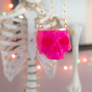 May include: A vibrant pink skull pendant hangs from a gold-colored chain. The skull is the focal point, with its hollow eye sockets and detailed features. The background includes a skeleton and string lights, suggesting a Halloween or gothic theme.