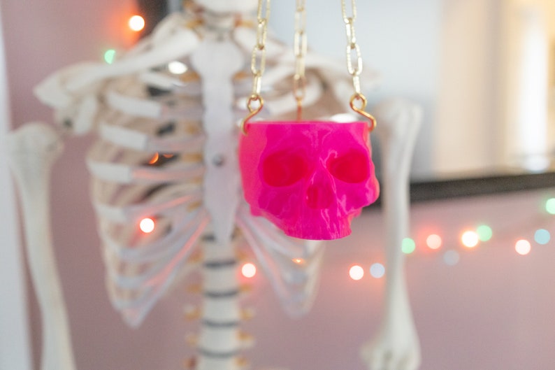 May include: A vibrant pink skull pendant hangs from a gold-colored chain. The skull is the focal point, with its hollow eye sockets and detailed features. The background includes a skeleton and string lights, suggesting a Halloween or gothic theme.