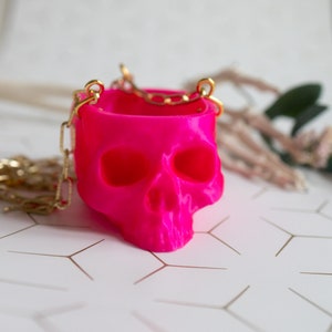 May include: A vibrant fuchsia skull-shaped container with a gold chain handle. The container has eye sockets and a nose cavity, and the chain is attached to the top of the skull. The background is a light color with a geometric pattern.