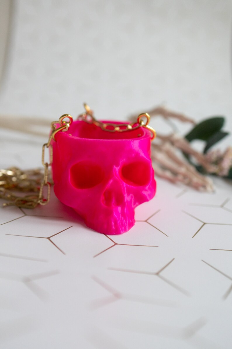 May include: A vibrant fuchsia skull-shaped container with a gold chain handle. The container has eye sockets and a nose cavity, and the chain is attached to the top of the skull. The background is a light color with a geometric pattern.