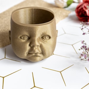 May include: A gold-coloured, 3D printed planter shaped like a baby's head. The planter has a smooth, textured surface and is sitting on a white background with a geometric pattern.