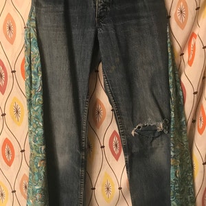 May include: Blue denim jeans with a distressed knee and floral fabric trim on the legs. The fabric trim is teal and purple.