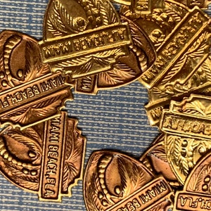 May include: Close-up of a group of gold-colored metal charms with the text "Miami Beach Fla" on them. The charms are shaped like a heart and have a palm tree design.