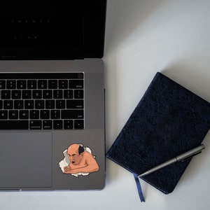 Peut inclure: Un autocollant repr&eacute;sentant un homme de dessin anim&eacute; avec une expression surprise, qui sort d'un ordinateur portable. L'autocollant est sur le pav&eacute; tactile de l'ordinateur portable argent&eacute;. Un carnet noir avec un stylo est sur la surface blanche &agrave; c&ocirc;t&eacute; de l'ordinateur portable.