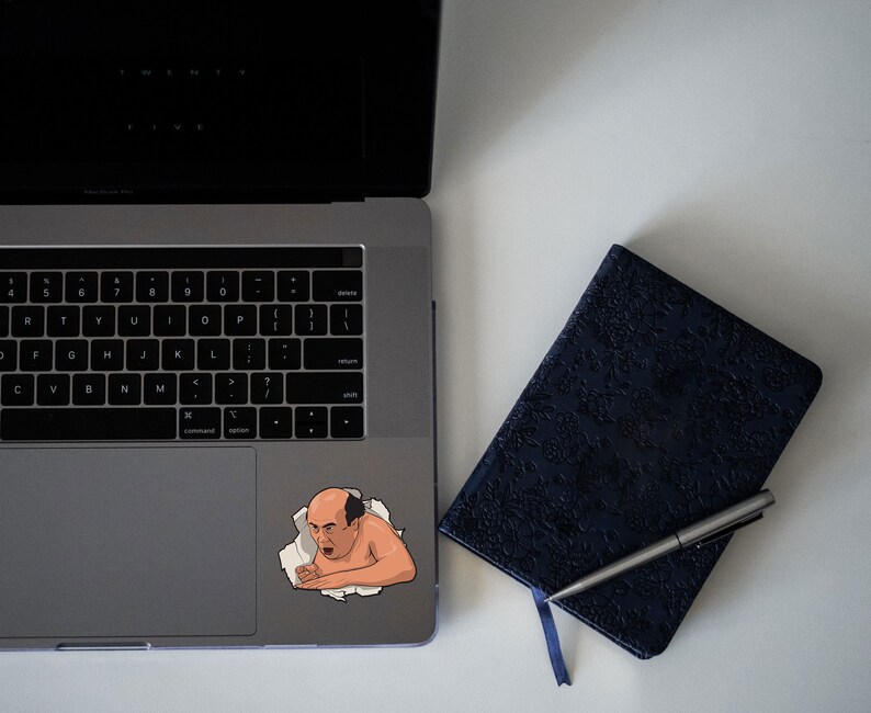 Peut inclure: Un autocollant repr&eacute;sentant un homme de dessin anim&eacute; avec une expression surprise, qui sort d'un ordinateur portable. L'autocollant est sur le pav&eacute; tactile de l'ordinateur portable argent&eacute;. Un carnet noir avec un stylo est sur la surface blanche &agrave; c&ocirc;t&eacute; de l'ordinateur portable.